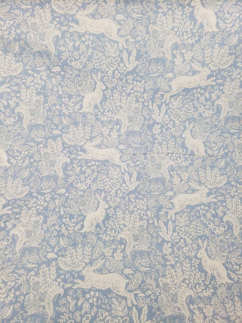 May include: Light blue fabric with a repeating pattern of white rabbits and foliage. The design features running and seated rabbits amidst detailed floral and plant elements, creating a whimsical, nature-inspired print. Ideal for home decor or crafting projects.
