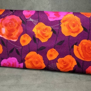 May include: A fabric bolt featuring a floral pattern with large orange and pink roses on a dark purple background. The design includes black stems, leaves, and white spiderwebs, creating a Halloween theme.