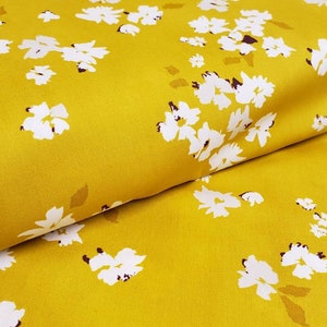 May include: Mustard yellow fabric with a repeating pattern of white floral blossoms and small brown accents. The fabric is folded, showing the texture and drape of the material. Ideal for sewing projects.