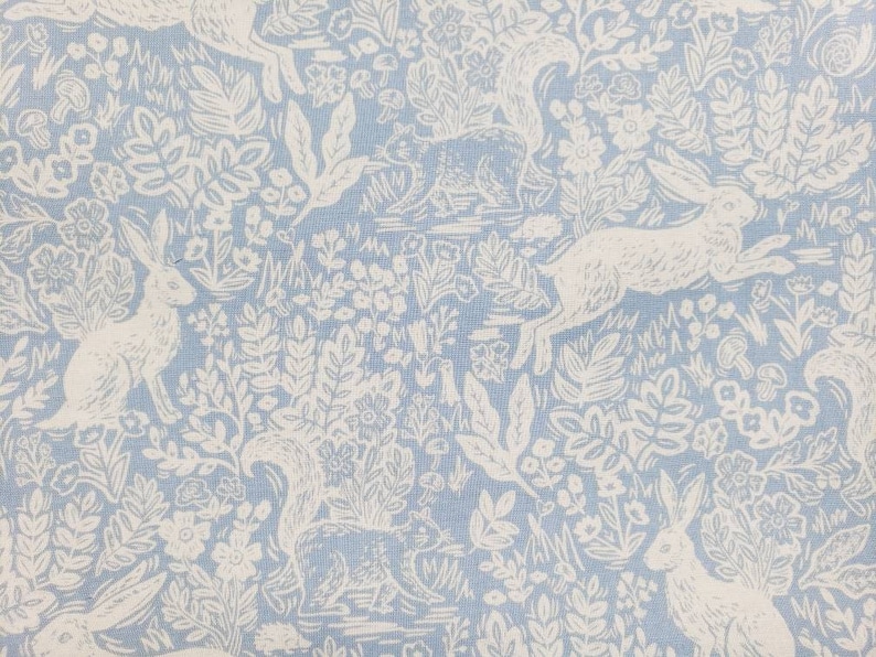 May include: Light blue fabric with a repeating pattern of white rabbits, squirrels, flowers, and foliage. The design has a vintage, storybook aesthetic, with detailed illustrations of woodland creatures and plants.