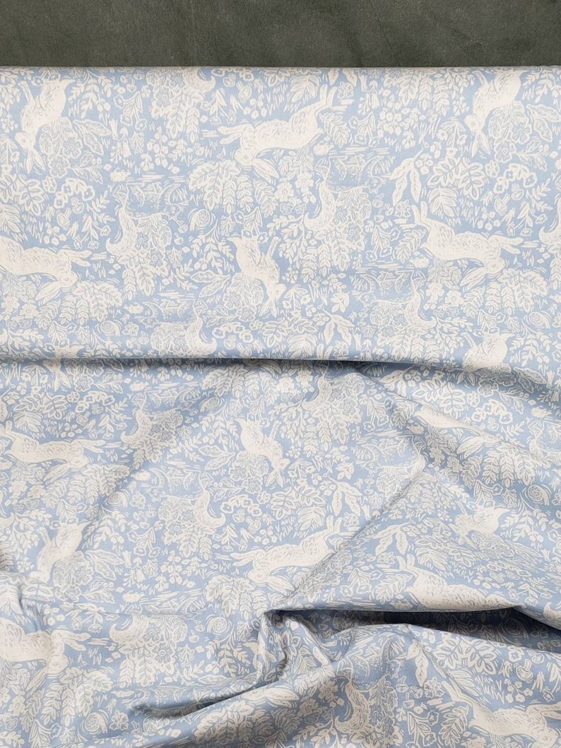 May include: Light blue fabric with a repeating pattern of white hares, birds, and foliage. The design is detailed and creates a whimsical, nature-inspired aesthetic. The fabric is folded, showing its texture and drape.