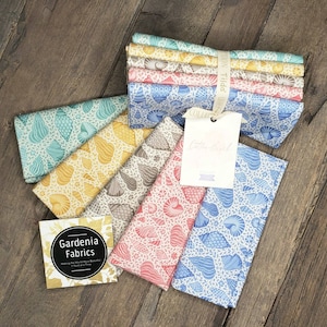 May include: A collection of folded cotton fabric pieces featuring various seashell patterns in shades of blue, yellow, pink, and beige. A white tag with the text Cotton Beach and a square card labeled Gardenia Fabrics are attached to the fabric bundle.