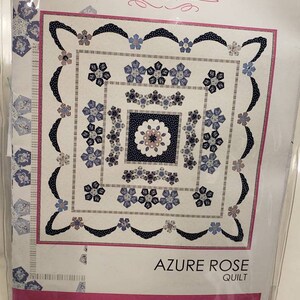 May include: A quilt pattern titled "Azure Rose" by Sue Daley Designs. The design features a central square with a floral pattern in shades of blue and white, surrounded by decorative borders and floral accents. The pattern is packaged in a clear plastic sleeve.
