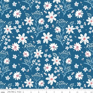 May include: A blue fabric featuring a repeating pattern of white flowers with pink centers, light blue leaves, and small white dots. A ruler at the bottom shows a scale of 12 inches, with the text Riley Blake Designs printed on the edge.
