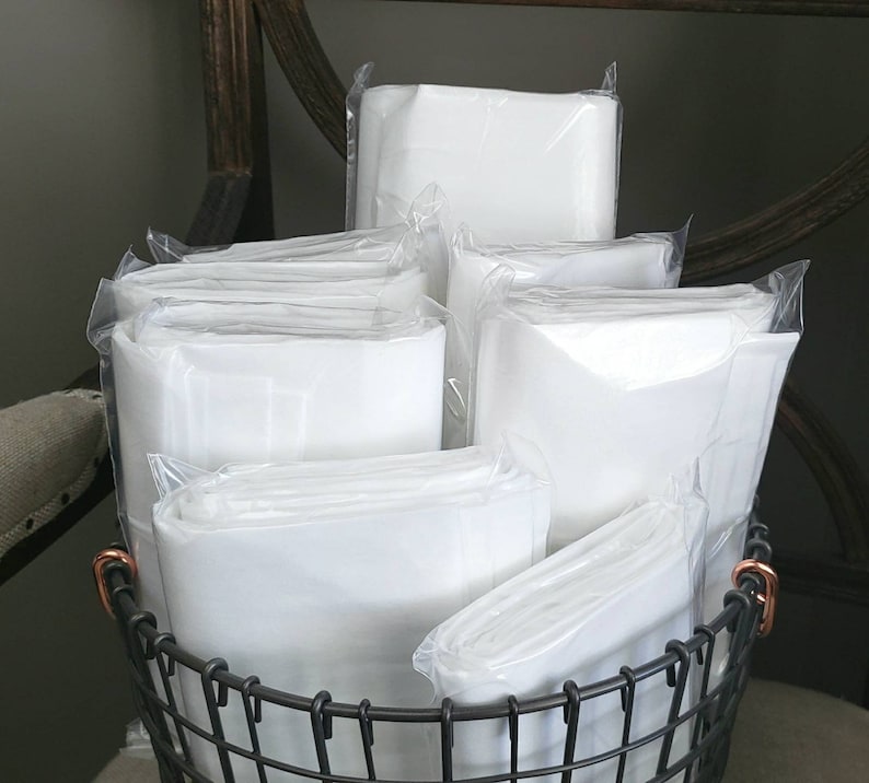 May include: A stack of white fabric squares wrapped in clear plastic, sitting in a black wire basket. The fabric squares are folded and stacked on top of each other.