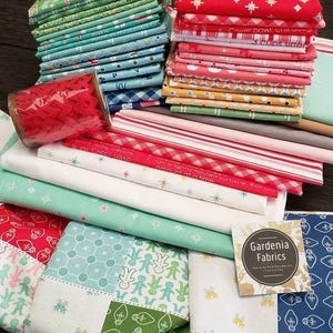 May include: A stack of colorful fabric squares with various holiday-themed patterns, including gingerbread men, snowflakes, and lights. The fabric is arranged in a pile with a red ribbon on top. The text "Gardenia Fabrics" is visible on a white tag.