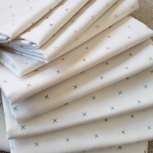 May include: A stack of ten white fabric squares with a repeating pattern of small crosses in various colors, including red, orange, green, and blue.