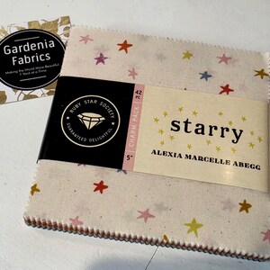 May include: A 42-piece 5-inch charm pack of fabric squares by Ruby Star Society titled Starry by Alexia Marcelle Abegg. The fabric features a cream background with small multi-colored stars in red, yellow, pink, and blue. A Gardenia Fabrics card is included.