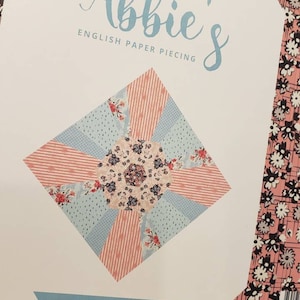 May include: A paper piecing pattern by Sue Daley Designs, featuring the text "Abbie's English Paper Piecing" and "Block No. 4." The design includes a central floral pattern with light blue, pink, and white accents. The background is a pink floral pattern.