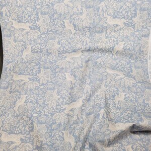 May include: Light blue fabric with a repeating pattern of white hares and foliage. The design features detailed illustrations of rabbits, flowers, and leaves, creating a whimsical and nature-inspired aesthetic. This fabric is suitable for various sewing and crafting projects.