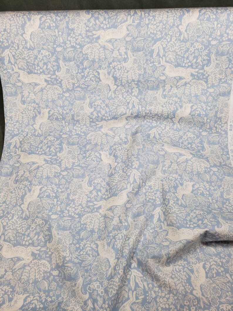 May include: Light blue fabric with a repeating pattern of white hares and foliage. The design features detailed illustrations of rabbits, flowers, and leaves, creating a whimsical and nature-inspired aesthetic. This fabric is suitable for various sewing and crafting projects.