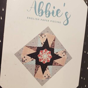 May include: A black and white box featuring a colorful star-shaped quilt block design. The text "Abbie's English Paper Piecing" is displayed in a script font. The box is labeled "Block No. 6" and includes the Sue Daley Designs logo.