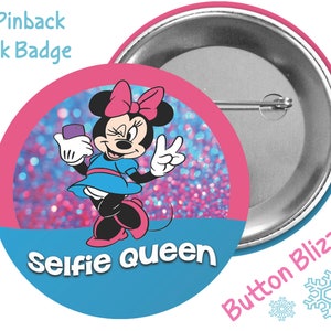 May include: A 3-inch pinback park badge featuring a cartoon image of Minnie Mouse wearing a blue dress and holding a smartphone. The background is pink and blue glitter with the text "Selfie Queen". The back of the badge is silver with the text "Button Blizzard" and snowflakes.