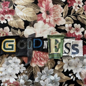 Op de afbeelding: Het woord GOODNESS gespeld met individuele stoffen lapjes met variërende kleuren, lettertypes en texturen, waaronder groene, zwarte, blauwe en witte letters, horizontaal gerangschikt tegen een zwarte bloemenstof achtergrond met roze en witte bloemen.