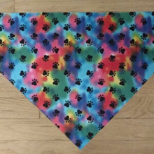 May include: A triangular bandana with a tie-dye pattern in shades of blue, green, red, and yellow. The bandana is covered in black paw prints.