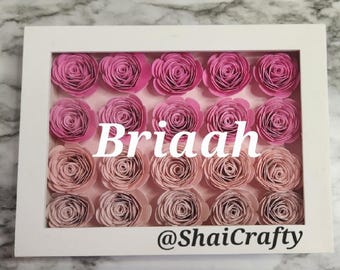 May include: A white rectangular shadow box frame containing twenty rolled paper roses arranged in four rows. The top two rows feature vibrant pink roses, while the bottom two rows feature muted mauve roses. The text Briaah and @ShaiCrafty are visible.