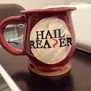 Pode incluir: Uma caneca de cerâmica bordô com uma alça grande. A caneca apresenta as palavras "HAIL REAPER" em letras pretas em um círculo creme. Uma caneta branca está apoiada em uma superfície escura ao lado da caneca.