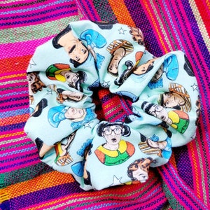 May include: A light blue scrunchie with a repeating pattern of cartoon faces. The faces are of various people with different hairstyles and expressions.