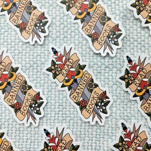 May include: A set of six stickers featuring a stylised dagger piercing through a banner that reads "Read Banned Books". The dagger is decorated with flowers and the banner is in a vintage style.