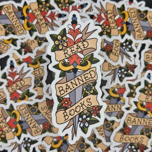 May include: A stack of stickers featuring a dagger design. The dagger has a red and white heart on the blade and is wrapped with a banner that reads "Read Banned Books". The stickers are white with black text and have a vintage tattoo style.