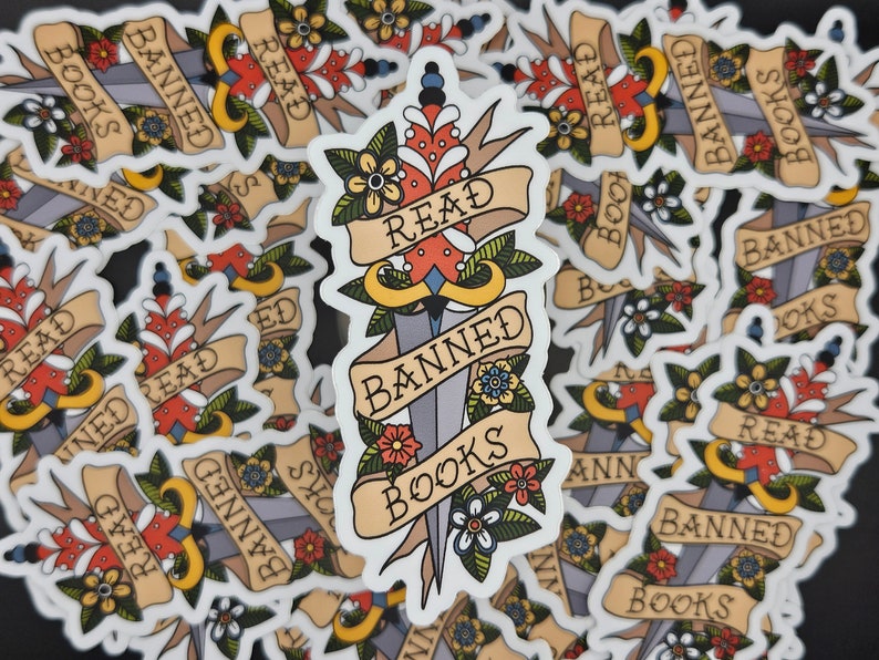 May include: A stack of stickers featuring a dagger design. The dagger has a red and white heart on the blade and is wrapped with a banner that reads "Read Banned Books". The stickers are white with black text and have a vintage tattoo style.