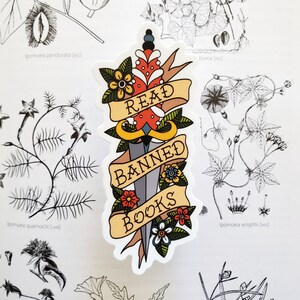 May include: A sticker featuring a dagger and a banner that reads "Read Banned Books". The dagger is adorned with red, yellow, and blue flowers. The banner is yellow with red and blue accents.