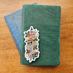 May include: A sticker featuring a stylised dagger with floral details. The dagger has a banner that reads "Read Banned Books".