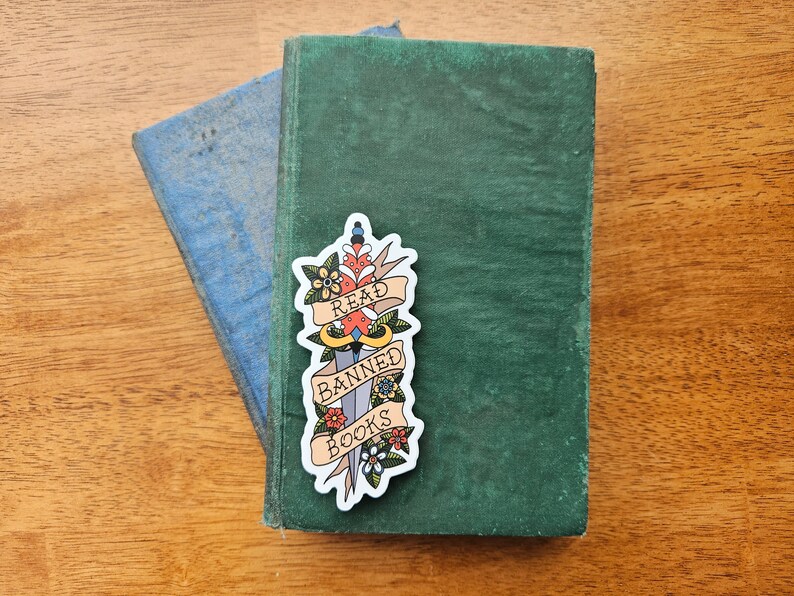 May include: A sticker featuring a stylised dagger with floral details. The dagger has a banner that reads "Read Banned Books".