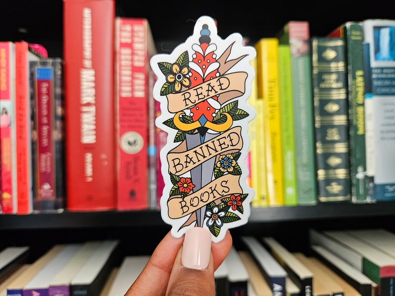 May include: A white sticker featuring a colourful tattoo-style illustration of a dagger with the words "Read Banned Books" on a banner. The sticker is being held in front of a bookshelf.