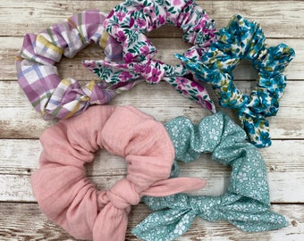 May include: Five fabric scrunchies with bows. The scrunchies are in various floral patterns, including pink, blue, and green.