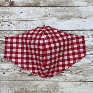 May include: A red and white gingham face mask with white elastic ear loops. The mask is made of fabric and has a classic checkered pattern. The mask is designed to cover the nose and mouth.