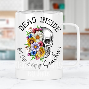 May include: White travel mug with a black skull with flowers and the text "Dead Inside But Still A Ray Of Sunshine".