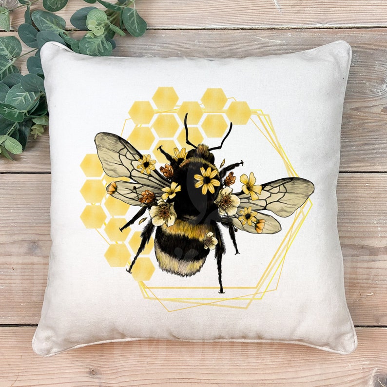 May include: A white pillow with a yellow and black bee illustration. The bee is surrounded by yellow flowers and a honeycomb pattern. The bee is in a gold hexagonal frame.