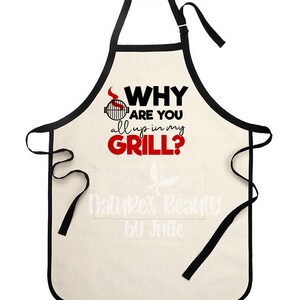 May include: A white cotton apron with black trim and adjustable straps. The apron has a large pocket on the front and features the text "WHY ARE YOU all up in my GRILL?" in black and red lettering.