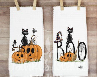 May include: Two white Halloween-themed kitchen towels hanging on a clothesline. One towel features a black cat, pumpkins, and a crow. The other towel says "BOO" with a witch hat, cat, pumpkins, and spider web.