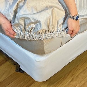 May include: A fitted bed sheet in a neutral beige color is being placed on a white bed frame. The sheet has an elasticized edge for a snug fit. The bed frame is white and sits on a wooden floor.