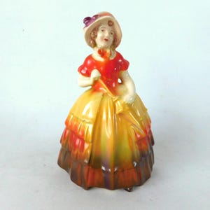 May include: A porcelain figurine of a woman in a red and yellow dress with a purple hat. The figurine is holding a yellow umbrella.