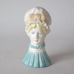May include: A vintage porcelain figurine of a woman's head and shoulders. The figurine features a white bonnet with a peach bow, blonde hair, and a light blue dress. The face has delicate features and is painted with soft colours.