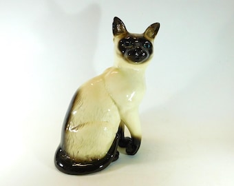 May include: A ceramic Siamese cat ornament. The cat is depicted in a seated position, with a cream-coloured body, dark brown markings on the face, ears, and tail, and blue eyes. The ornament has a glossy finish and is set against a white background.