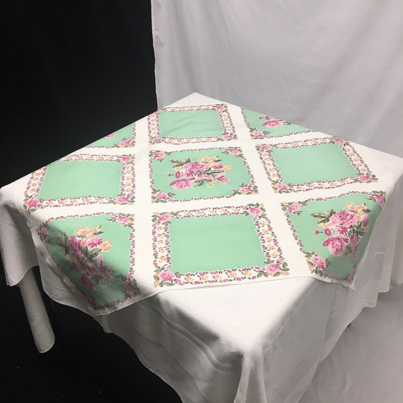 Op de afbeelding: Een wit tafelkleed met een groen en roze bloemenpatroon. Het tafelkleed heeft een vierkant ontwerp met een rand van roze en gele bloemen. Het midden van elk vierkant is effen groen.