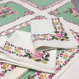 Op de afbeelding: Een set van vier witte linnen servetten met een bloemmotief in roze, gele en groene tinten. De servetten zijn gevouwen en gestapeld op een groene tafelkleed met een vergelijkbaar bloemmotief.