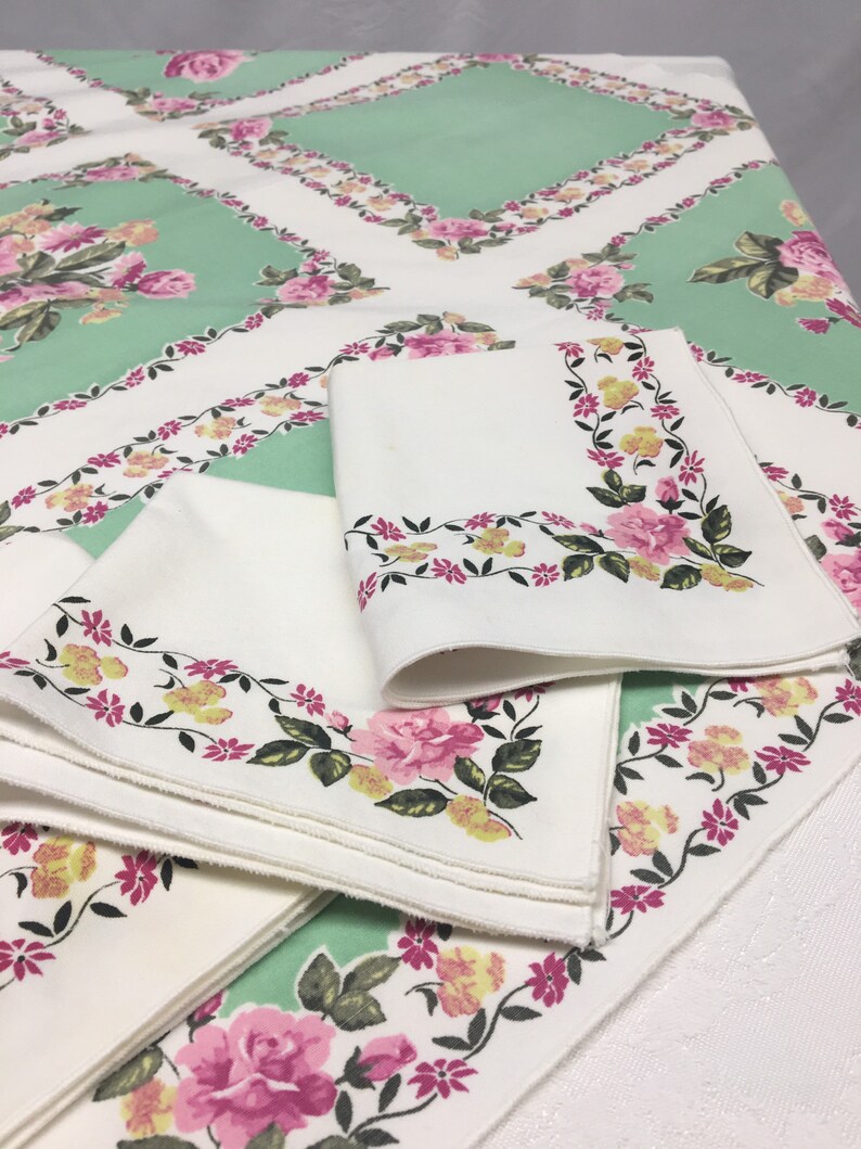 Op de afbeelding: Een set van vier witte linnen servetten met een bloemmotief in roze, gele en groene tinten. De servetten zijn gevouwen en gestapeld op een groene tafelkleed met een vergelijkbaar bloemmotief.