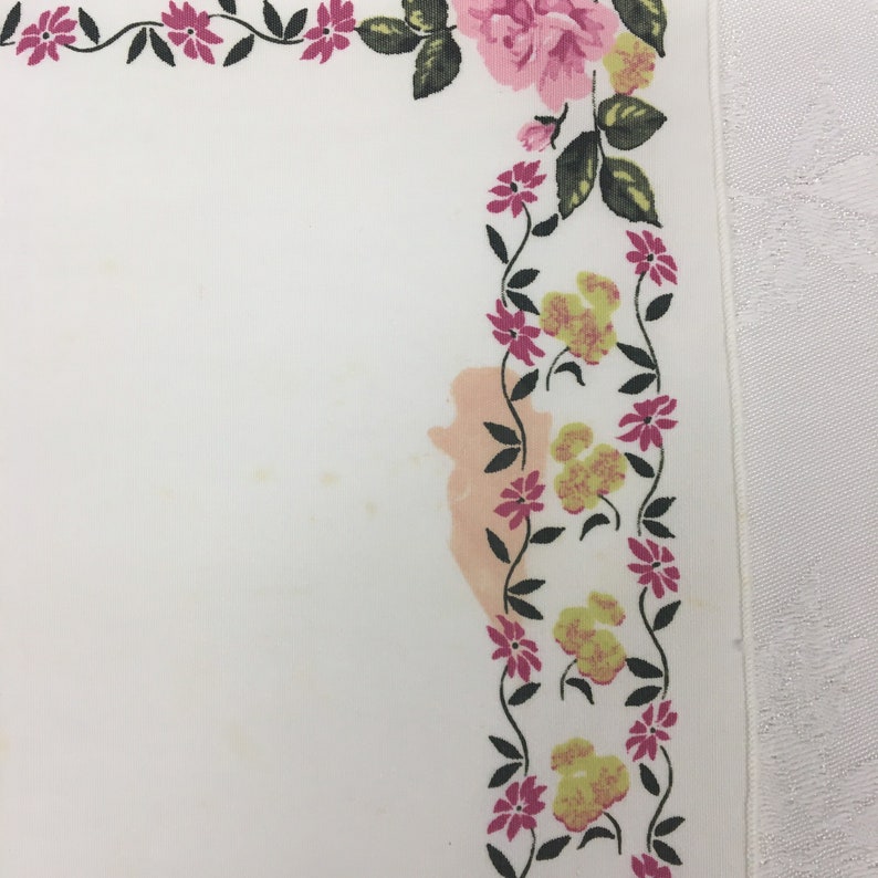 Op de afbeelding: Witte tafelkleed met een roze en gele bloemenrand. Het tafelkleed heeft een vlek in het midden.