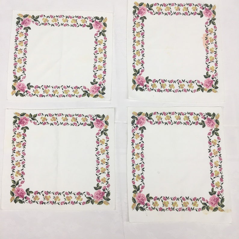 Op de afbeelding: Vier witte servetten met een roze en gele bloemenrand. De servetten zijn gevouwen en in een vierkant patroon gerangschikt.
