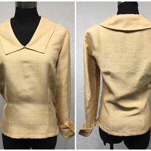 May include: A long-sleeved, light beige blouse with a wide, pointed collar. The blouse has a fitted silhouette and button cuffs.