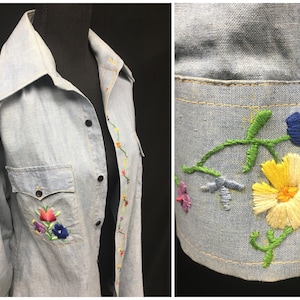 May include: A light blue denim shirt with embroidered flowers in various colors, including red, yellow, blue, and green. The shirt has a pointed collar, long sleeves, and a button-up front.