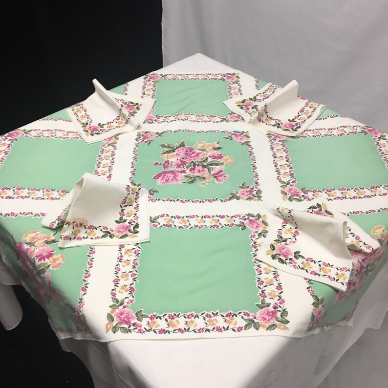 Op de afbeelding: Een groene tafelkleed met een bloemenpatroon en witte randen. Het tafelkleed is gedekt met zes witte servetten, elk gevouwen in een driehoek.