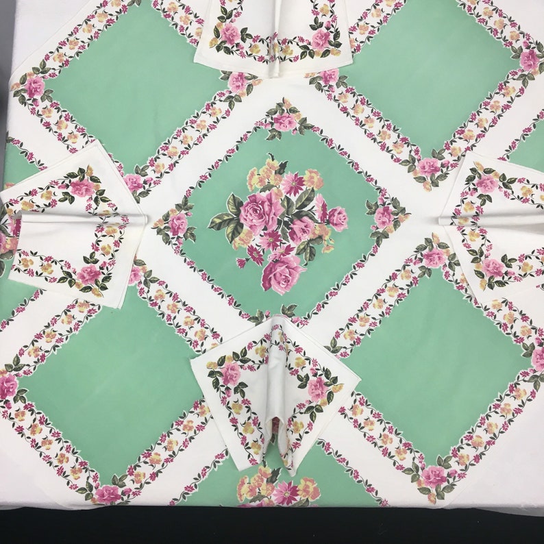 Op de afbeelding: Een wit tafelkleed met een groen en bloemig patroon. Het tafelkleed heeft een ruitpatroon met een bloemig ontwerp in het midden van elke ruit. Het tafelkleed is versierd met roze en gele bloemen.