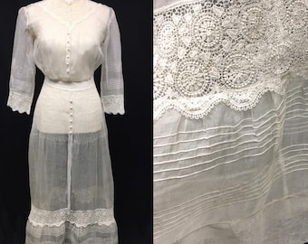 May include: A vintage white dress with a sheer skirt and long sleeves. The dress features intricate lace detailing on the bodice, sleeves, and skirt hem. The bodice has a button-down front. The dress is displayed on a mannequin against a black background.