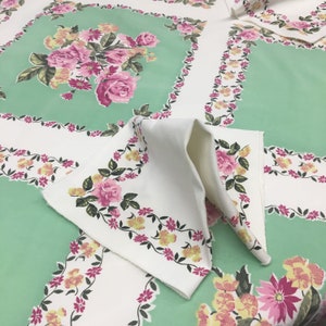 Op de afbeelding: Een wit tafelkleed met een groen bloemenpatroon. Het tafelkleed heeft een rand van roze en gele bloemen. Het tafelkleed is dubbelgevouwen en het bloemenpatroon is aan beide kanten zichtbaar.